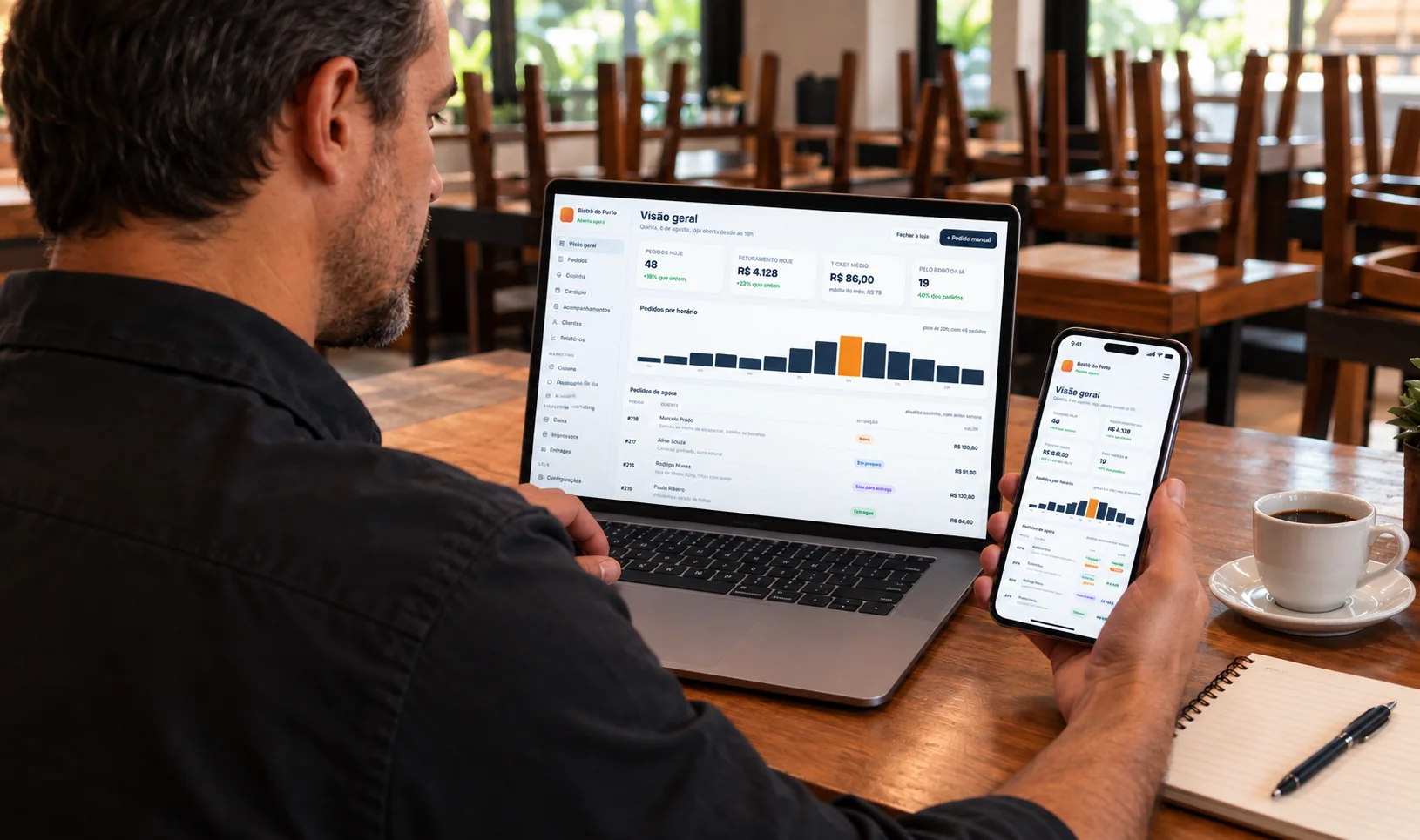 Dono do restaurante conferindo o painel no notebook e no celular, com o salão vazio ao fundo